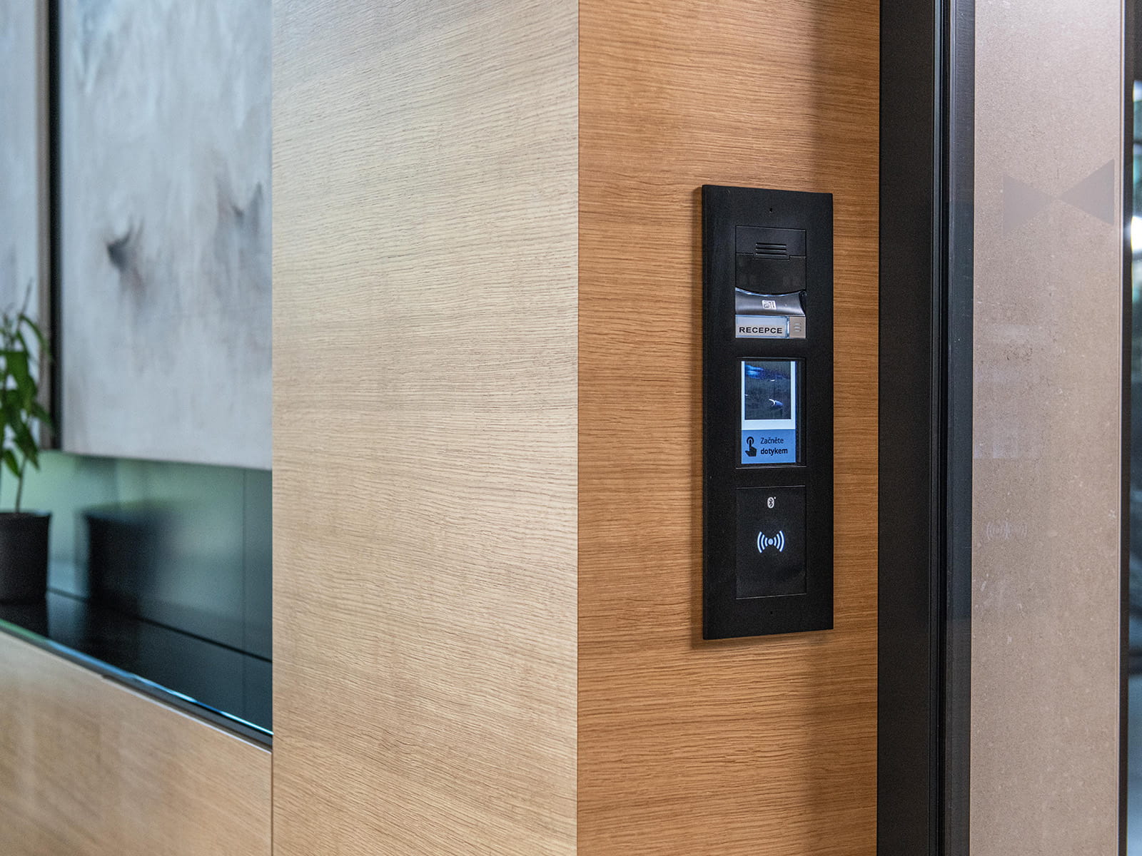 Panneau intercom noir monté au mur avec un écran et des boutons installé sur un mur en bois près d'une porte en verre.