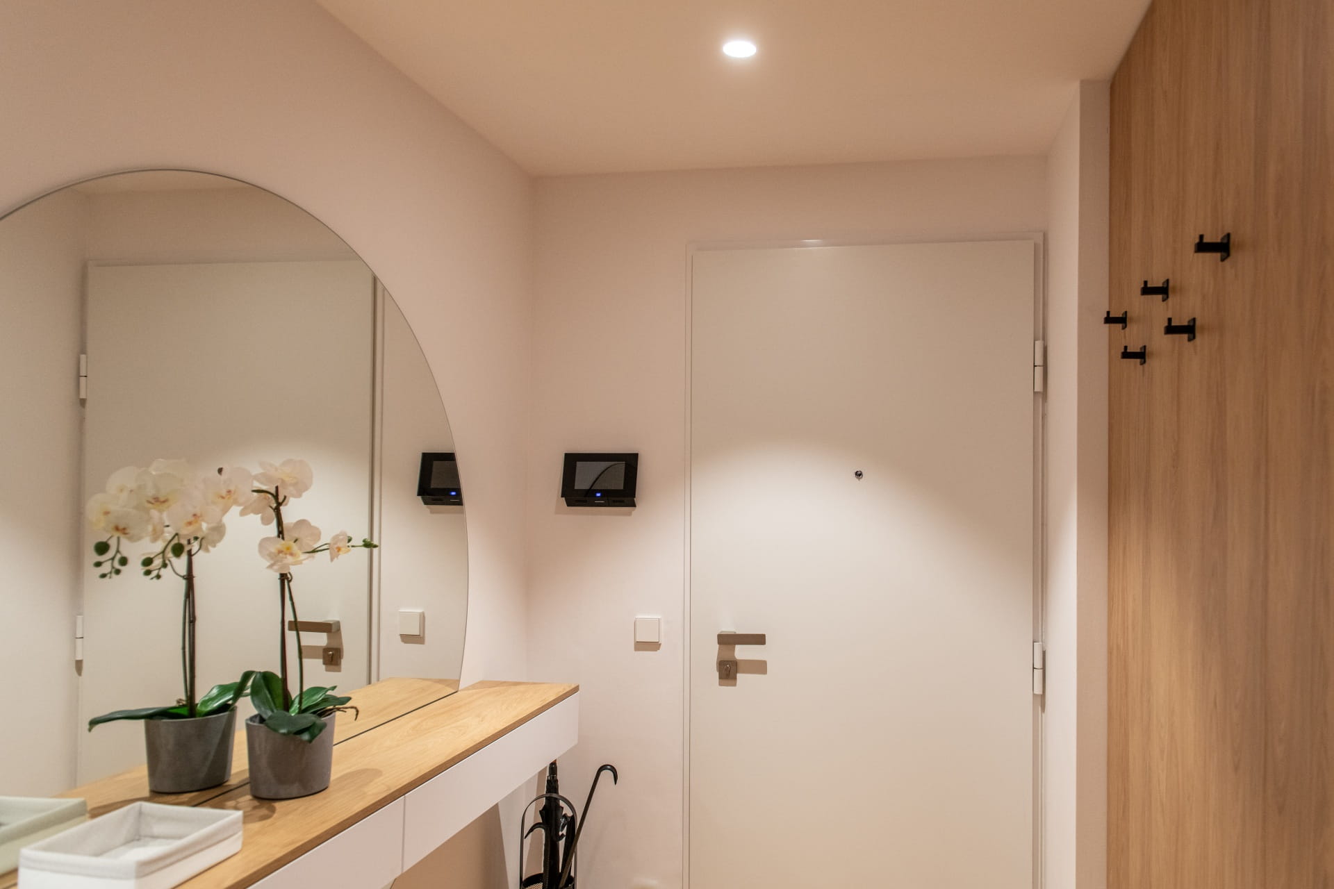Couloir moderne avec une porte d'entrée blanche, des accents en bois, un interphone mural et des orchidées décoratives sur une table console.