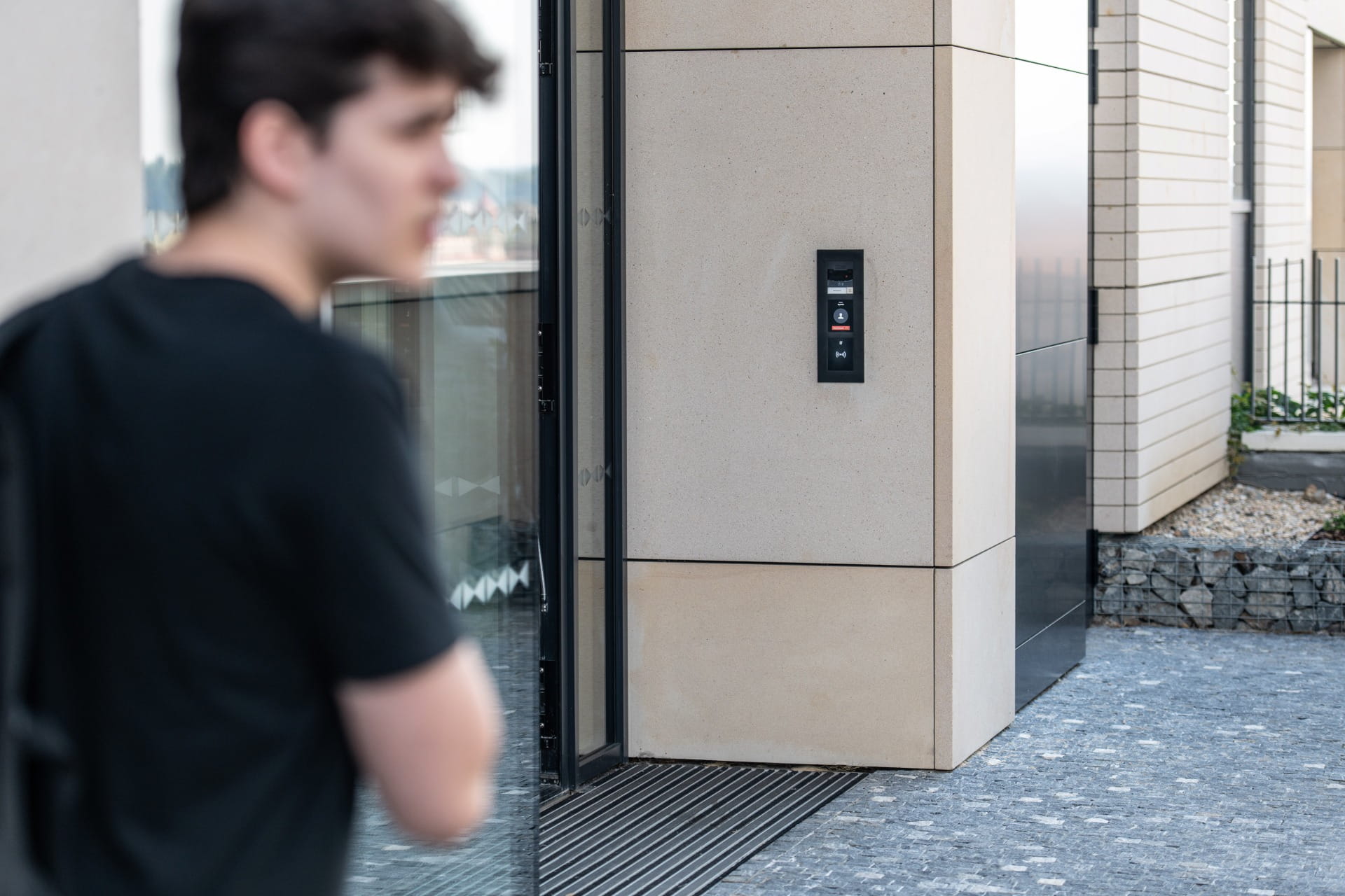 Personne se tenant près de l'entrée d'un bâtiment moderne avec un panneau d'interphone mural à côté d'une porte en verre.