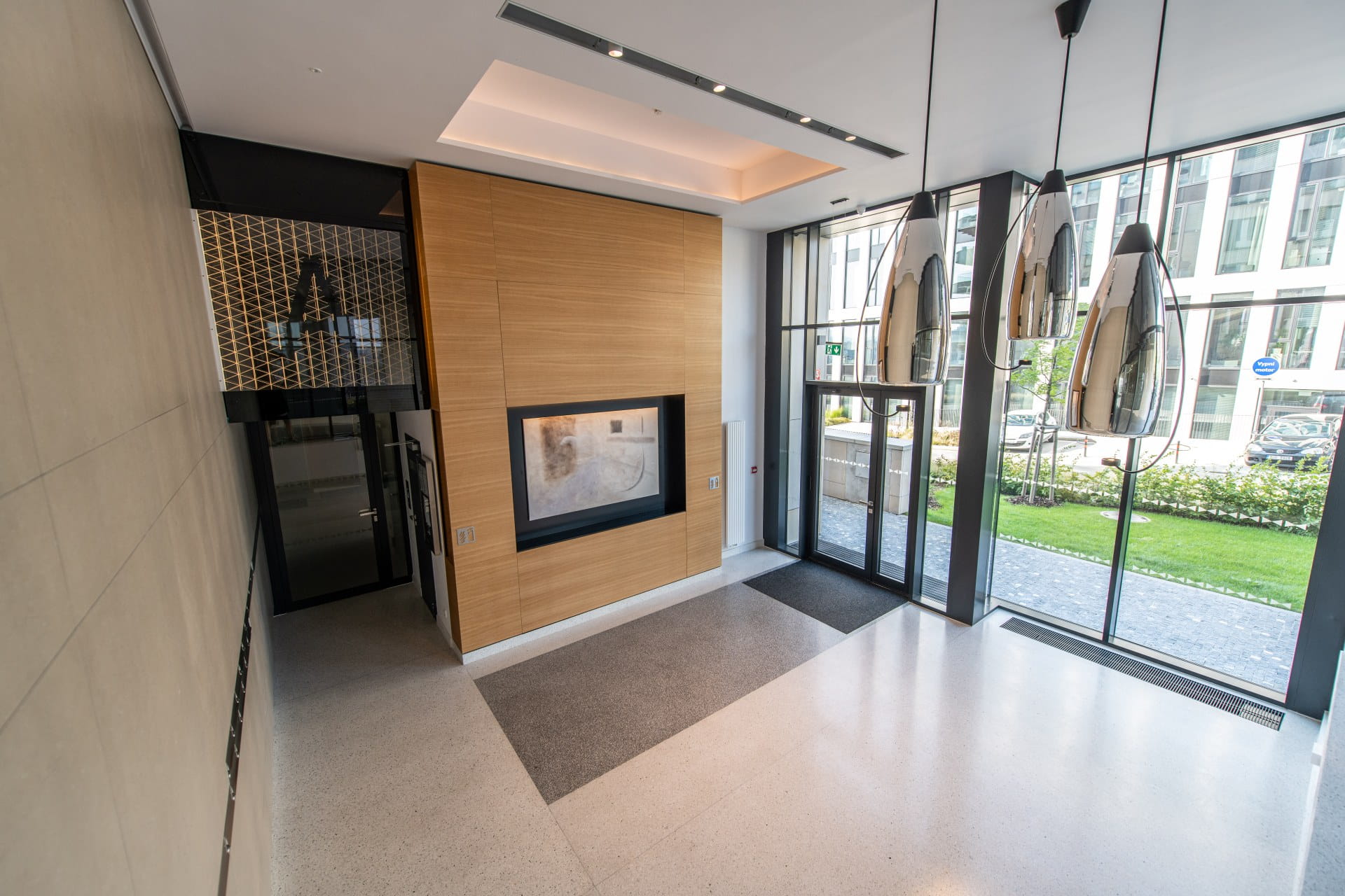 Hall d'entrée d'un bâtiment moderne avec de grandes portes en verre, un mur d'accent en bois, une entrée d'ascenseur et des lumières suspendues.