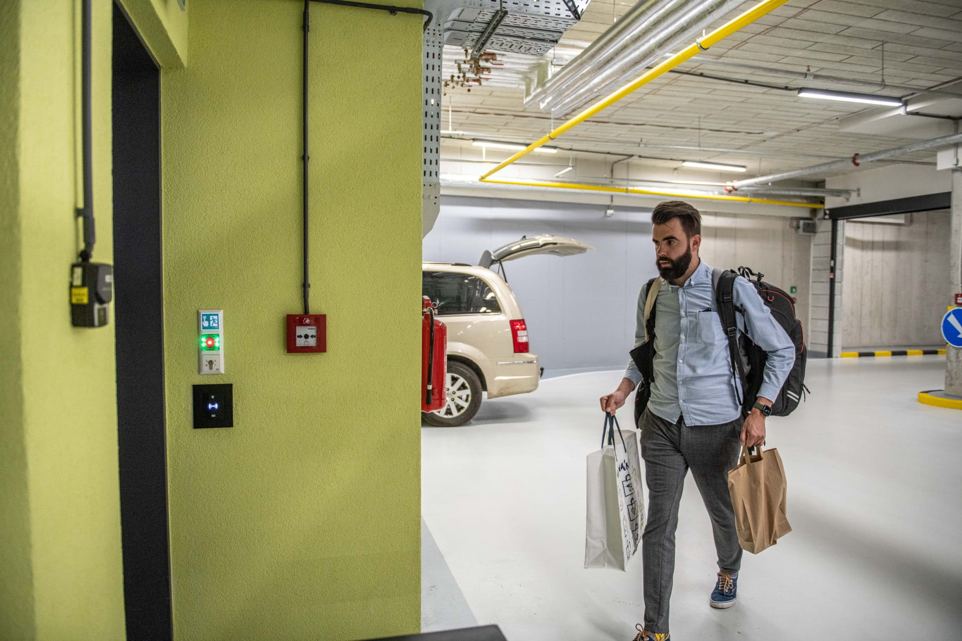 Personne marchant dans un garage souterrain portant des sacs de shopping, près d'un panneau de contrôle d'accès mural.