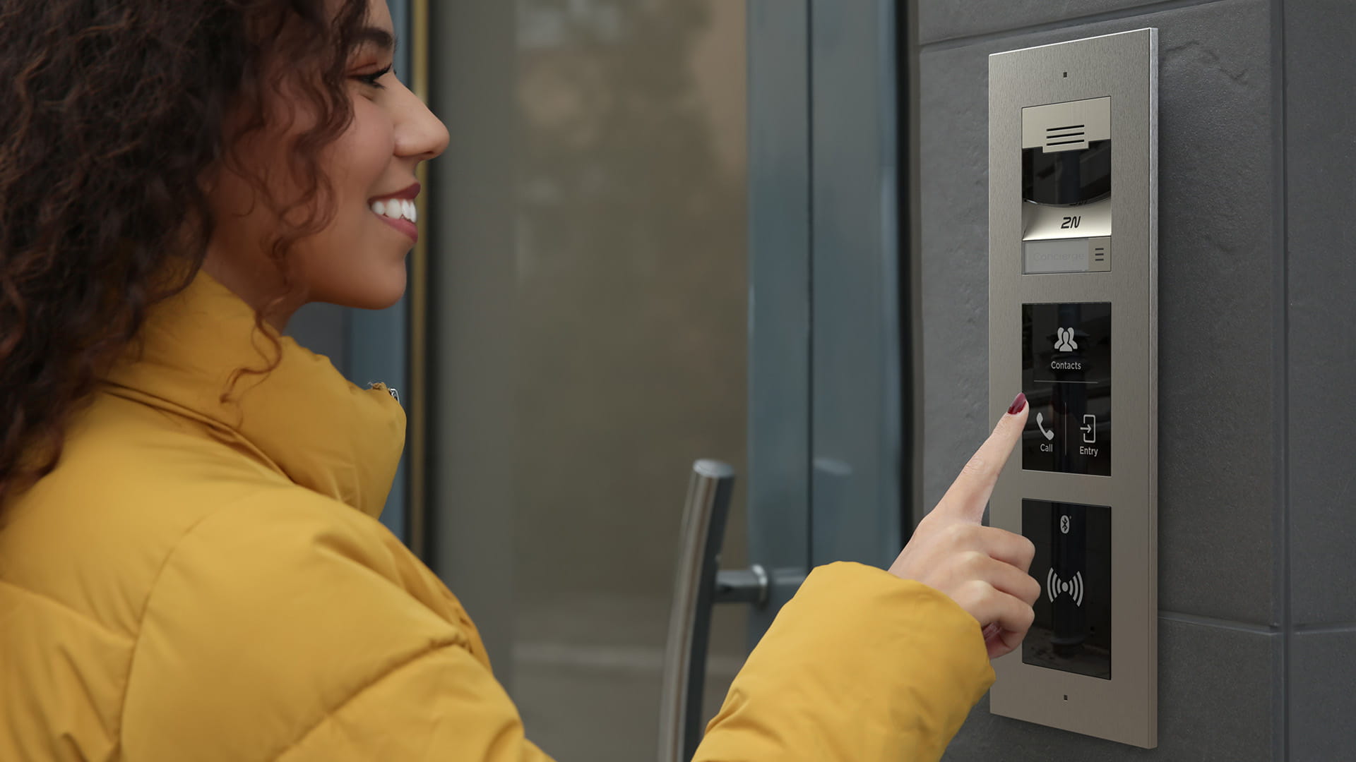 Person wearing a yellow jacket pressing a button on a stainless steel intercom panel mounted on a gray door frame.