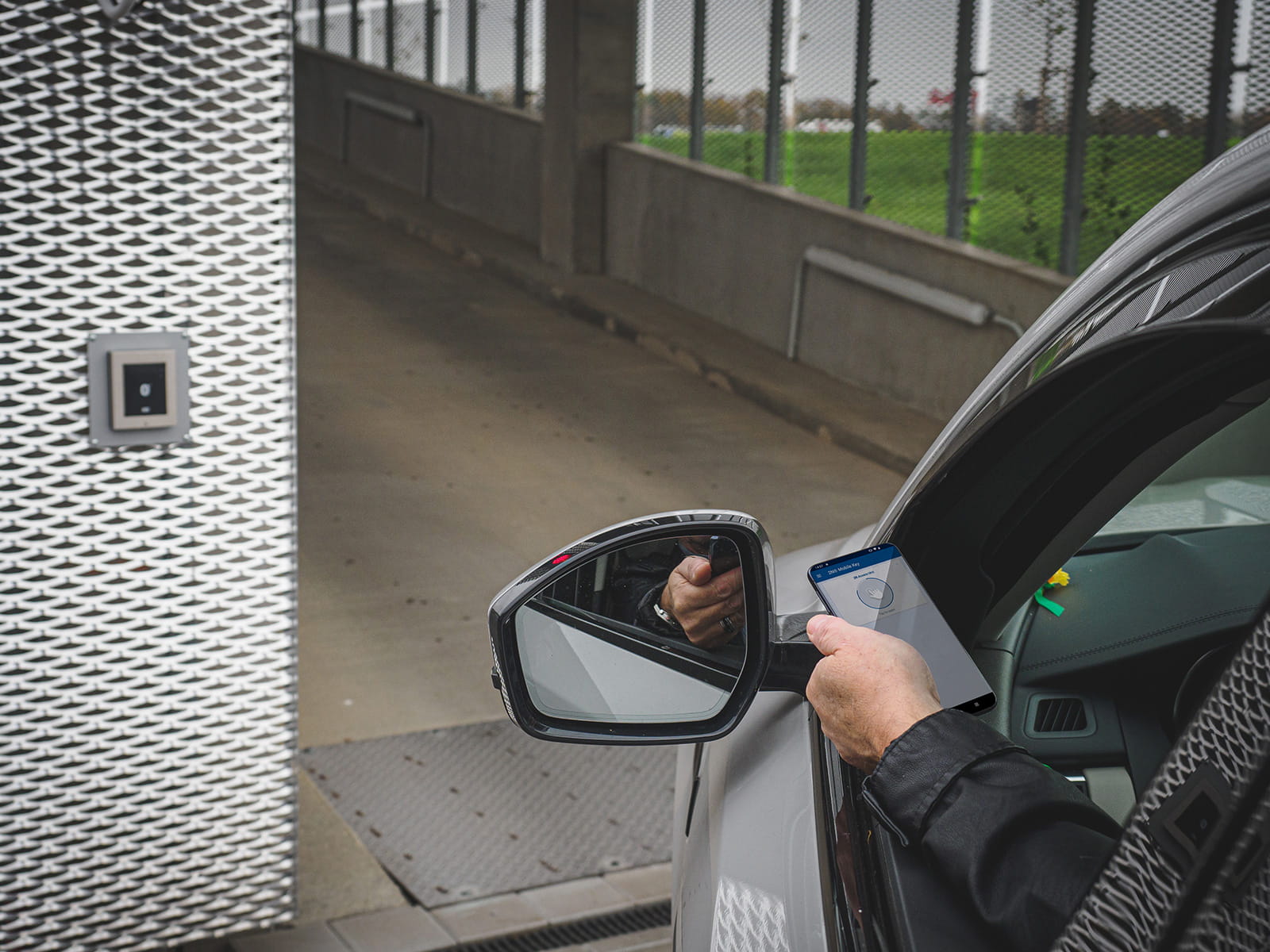 Osoba w samochodzie trzymająca smartfona w pobliżu zewnętrznego panelu dostępu przy bramie.