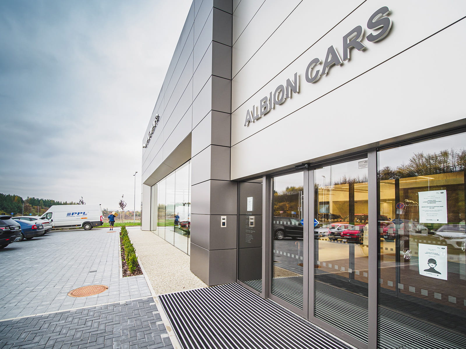 Nowoczesny budynek salonu samochodowego z dużymi szklanymi drzwiami i widocznym napisem „ARON CARS”.
