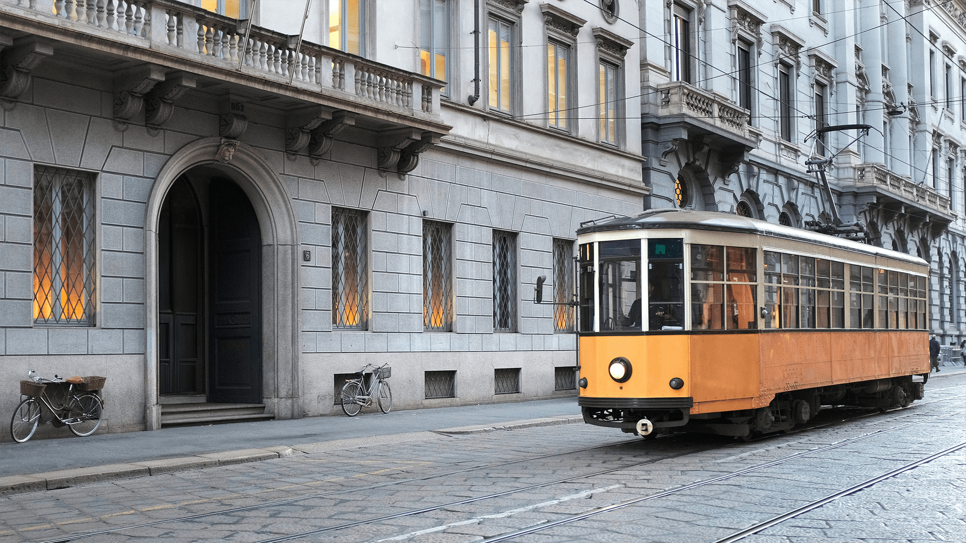 Un tramway vintage orange roule sur une rue pavée, passant devant des bâtiments historiques en pierre avec des vélos garés à côté.