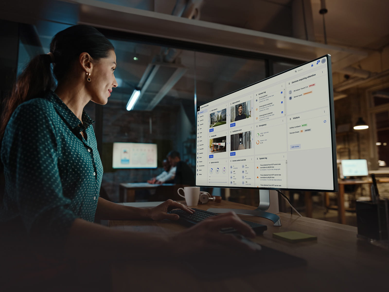 Person working at a desk using a large monitor displaying a security management interface with camera feeds and access logs.