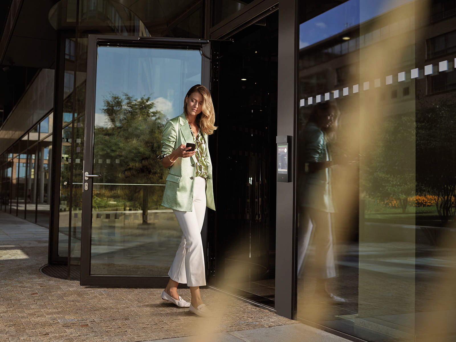 Person walking out of a modern glass building holding a smartphone near an open door.