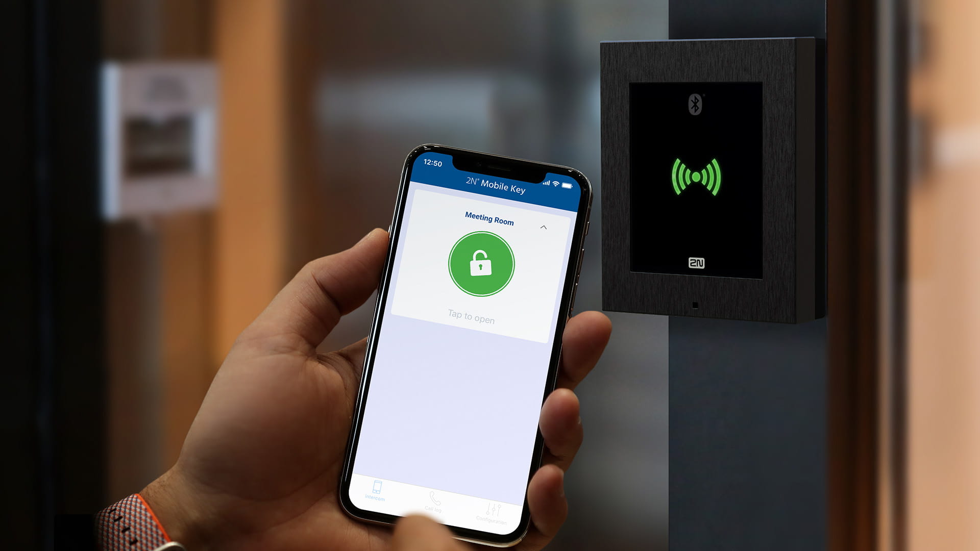 壁に取り付けられたRFIDリーダーの近くで、緑のアイコンを表示しているモバイルアクセスアプリを持ったスマートフォンを手に持っている。