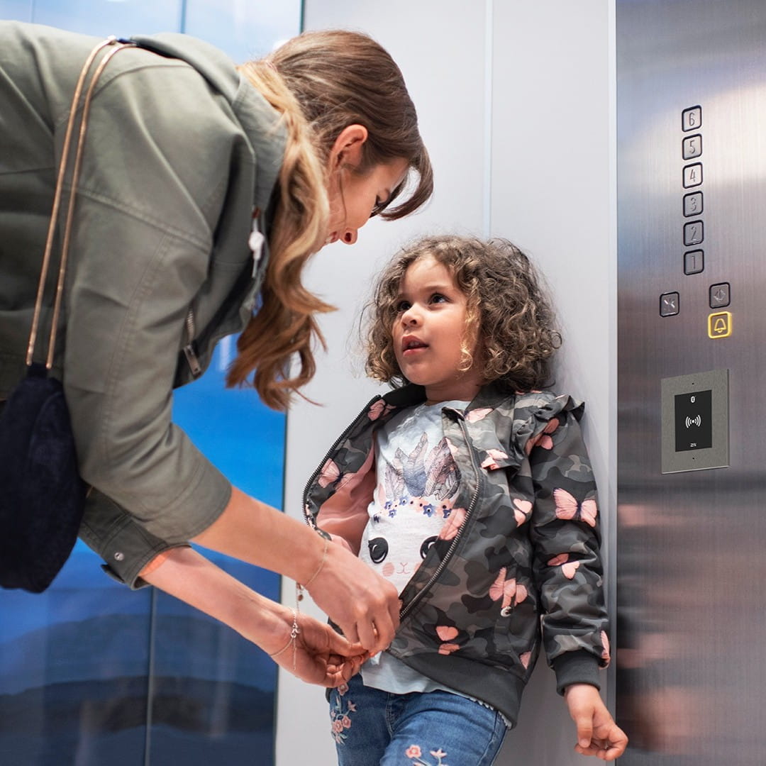 Volwassene helpt een kind in een lift, met een zichtbaar bedieningspaneel en metalen wanden op de achtergrond.