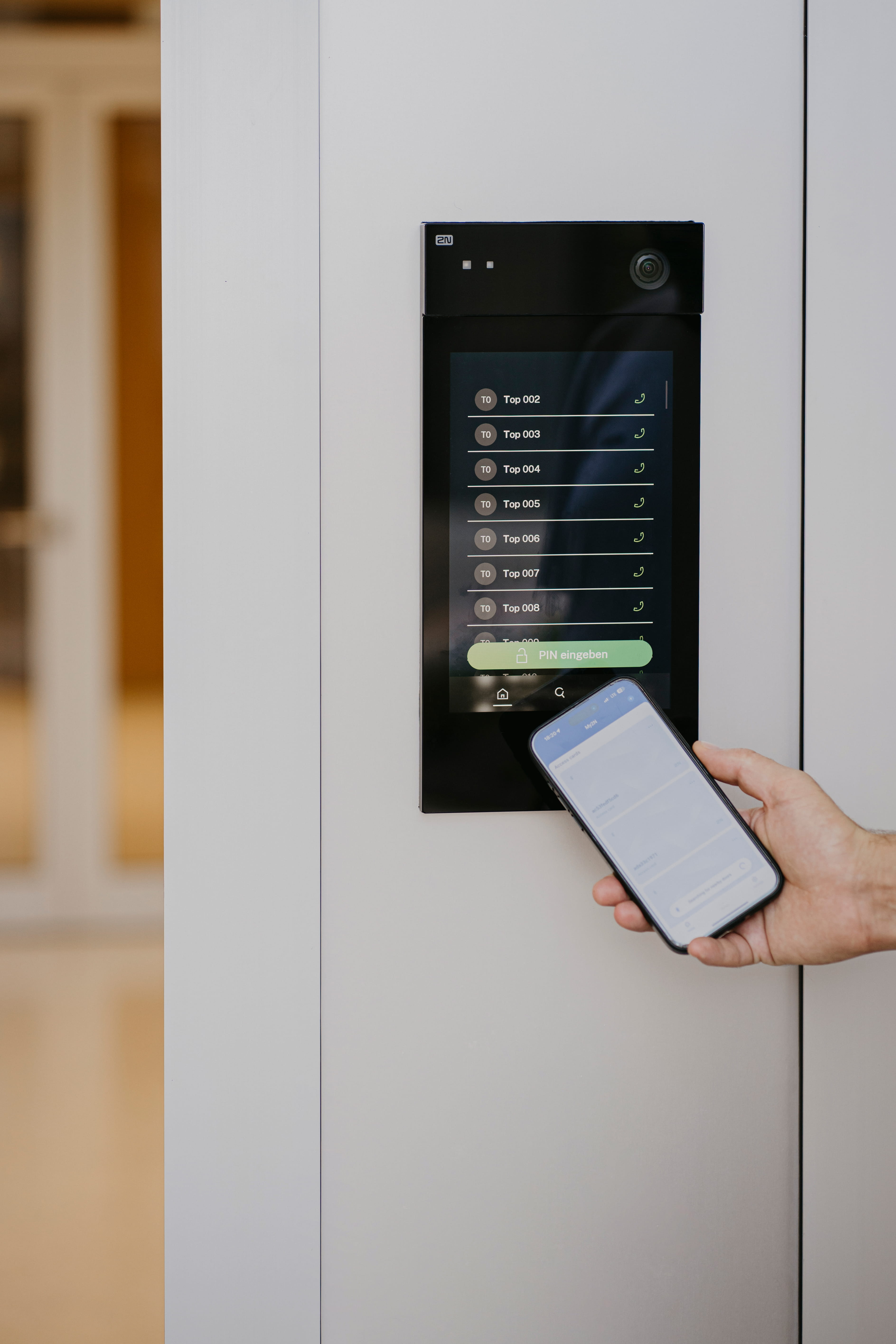 Main tenant un smartphone près d'un panneau d'accès à écran tactile monté sur le mur pour une entrée sécurisée.