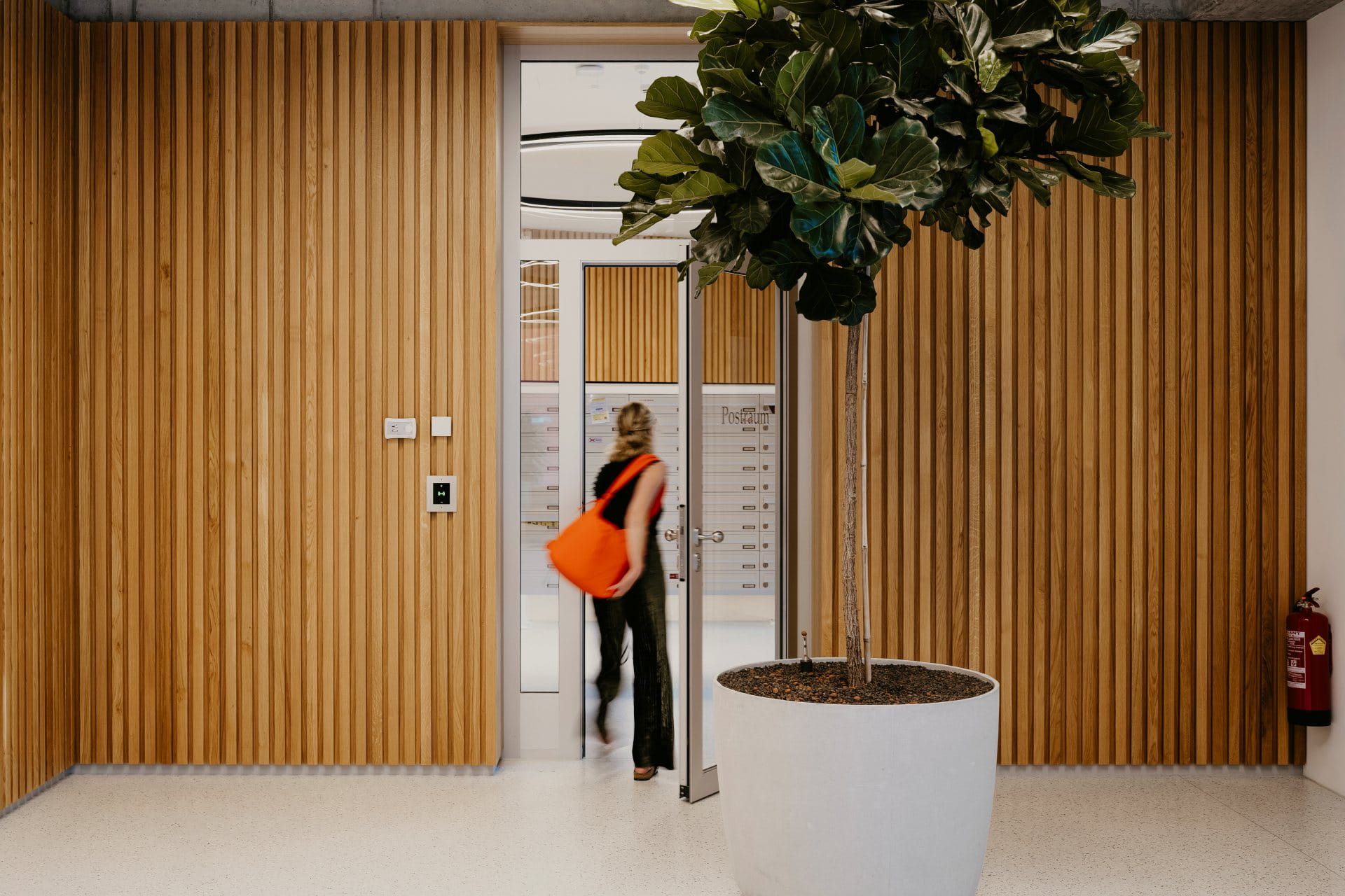 Personne avec un sac orange entrant dans un hall moderne avec des murs en panneaux de bois et une grande plante en pot.