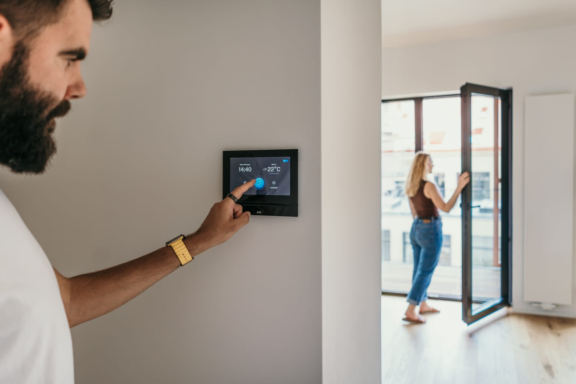 Personne utilisant un panneau de contrôle à écran tactile monté sur le mur pendant qu'une autre personne se tient près d'une porte en verre ouverte.