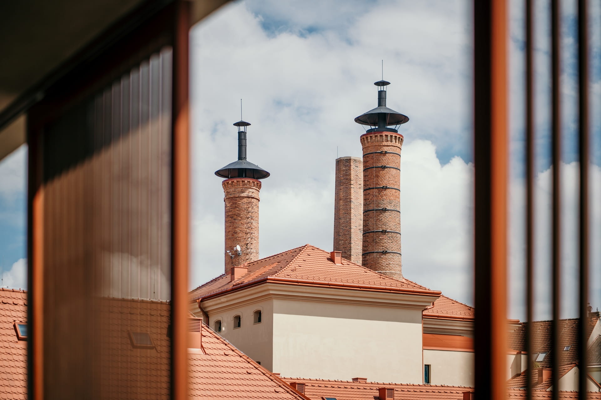 Vue de cheminées en briques historiques et de toits en tuiles rouges encadrés par des panneaux de verre modernes sous un ciel nuageux.