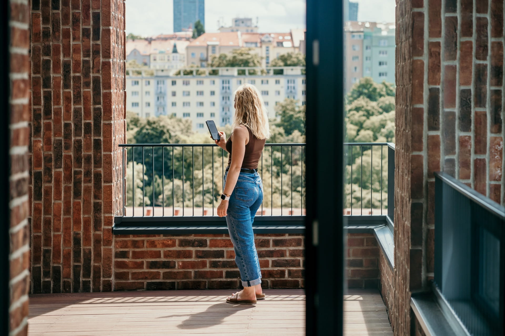 Une personne debout sur un balcon en briques, tenant un smartphone, avec une vue sur la ville en arrière-plan.
