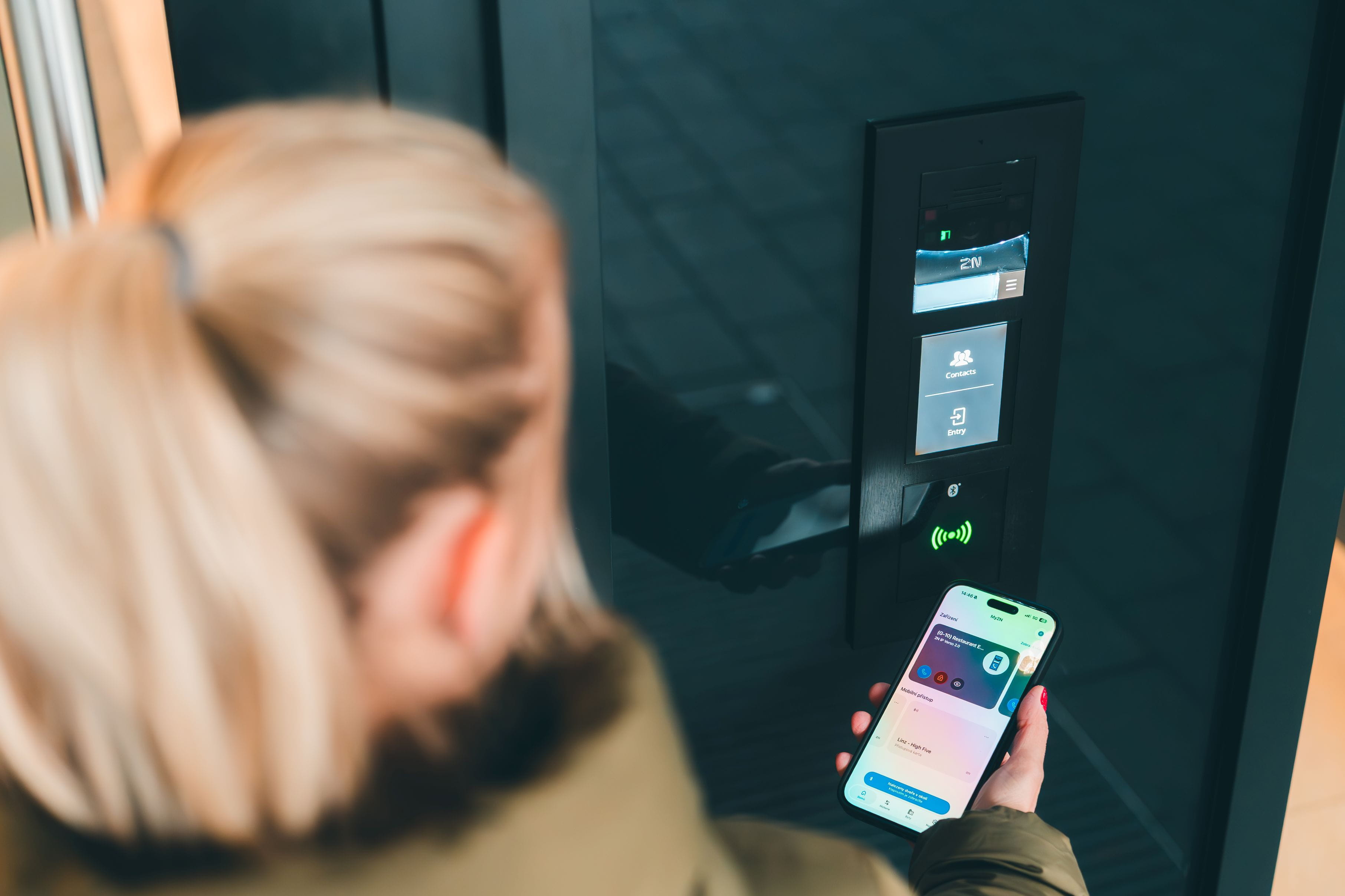 Persoon die een smartphone vasthoudt met een toegang-app dicht bij een 2N-deur apparaat dat een groene indicator toont.