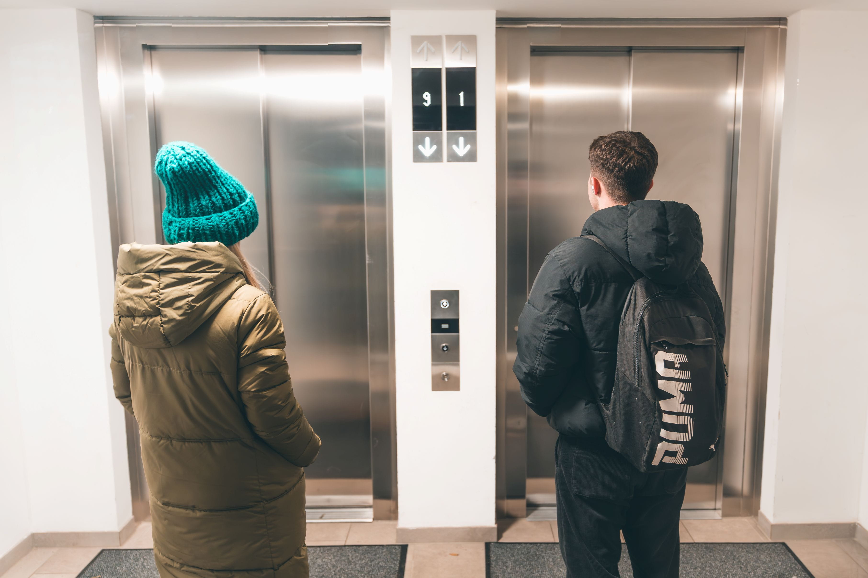 Twee mensen staan voor twee gesloten liftdeuren in een helder verlichte gang.