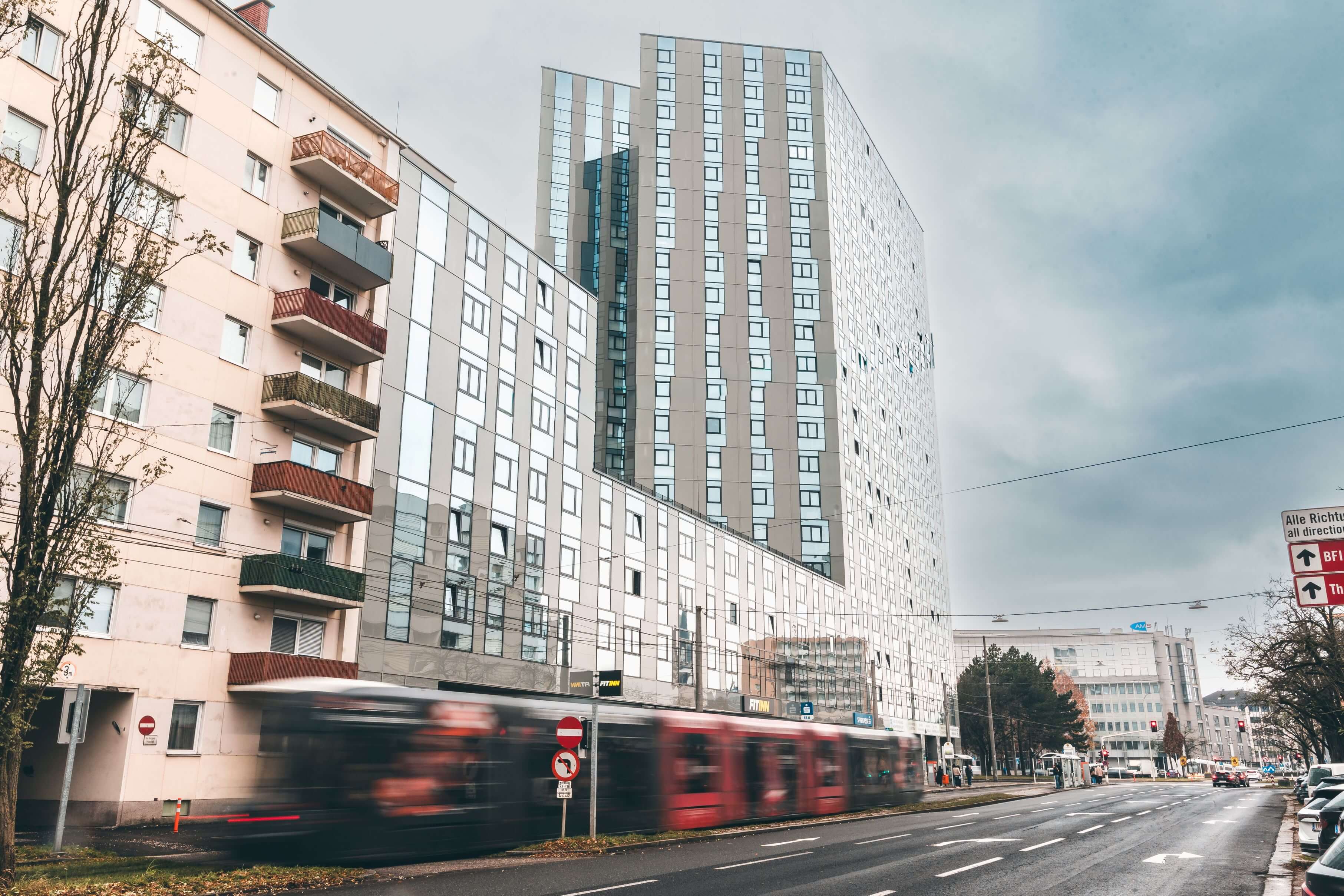 Rue de la ville avec un grand bâtiment moderne en verre, des appartements plus anciens et un tram flou qui passe.