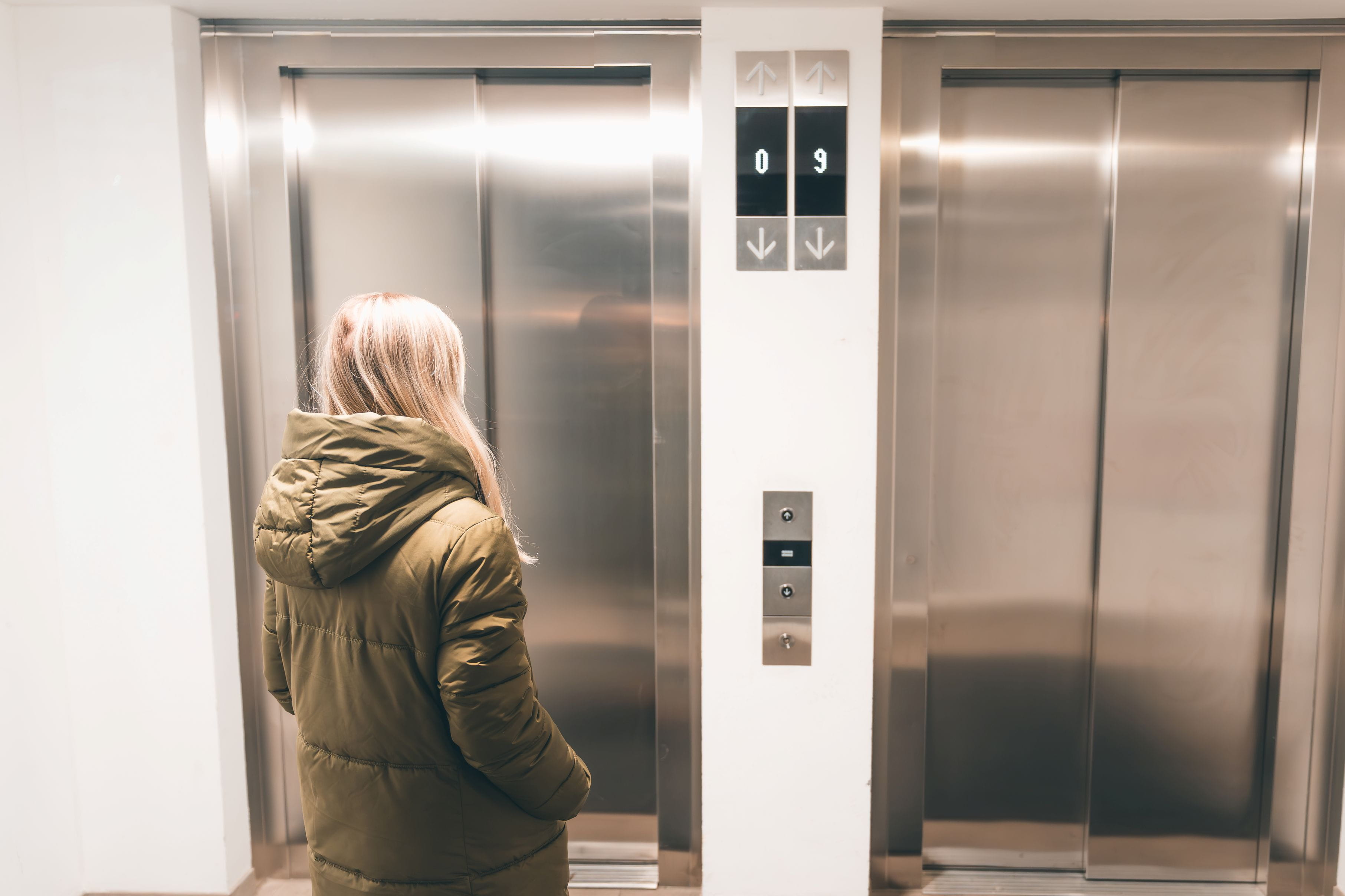 Persoon met een rugzak die voor twee gesloten liftdeuren staat en een klein pakket vasthoudt.